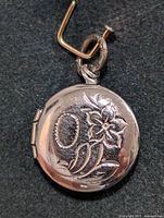 Front view of the silver round locket pendant showing detailed flower and leaf engravings with a loop for chain attachment.