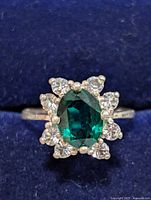Top view of silver ring showing oval green stone surrounded by clear stones on a blue background.