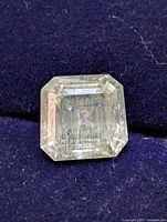 Close-up image of a transparent square-shaped Asscher cut gemstone on a dark fabric background, showing internal inclusions and visible facet lines.