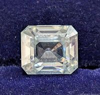Front view of rectangular emerald-cut clear gemstone resembling a diamond, showing its faceting and brilliance against a dark blue textured background.