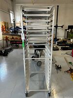 Front view of the bakers rack showing multiple slots with several metal trays inserted and caster wheels at the bottom.
