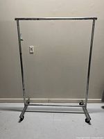 Front view of metal clothes rack on casters against wall, showing rectangular frame and top horizontal bar with extensions.