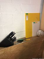 Photo showing one black knife block with knives, two cutting boards (yellow large, white small), and metal fluted mold on wooden surface against white cinderblock wall.