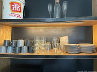 Shelf view showing stacked ceramic mugs, clear glasses, bowls, plates, and side plates.