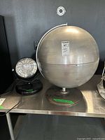 Front view of large silver metal globe with label and industrial light beside it on metal table, showing size and condition