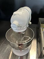 Front-angle view of white tilt-head mixer on metal surface showing bowl and attachments