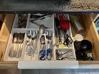Drawer with organized compartments containing stainless steel forks, knives, spoons, metal tongs, scissors, and other miscellaneous small kitchen utensils.