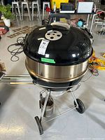 Front view of Weber charcoal barbecue grill with chimney starter attached underneath, black lid with vent holes and handle visible.
