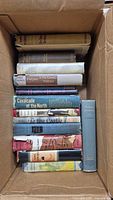 Top-down view of 13 vintage hardcover books in a cardboard box showing spines and some dust jackets, titles mostly legible.