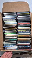 Top view of a cardboard box filled with many jewel case audio CDs, stacked vertically and horizontally, showing spines and covers.