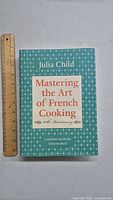 Front cover of the 50th anniversary edition of 'Mastering the Art of French Cooking' by Julia Child next to a ruler for size reference.