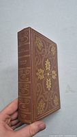 Front angled view showing the decorative gold embossed design on brown hardcover and book title and author on spine.
