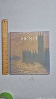 Front cover of the Monet art book showing impressionist painting with sunset and silhouette of building by water. A wooden ruler placed for size reference approx 10 inches tall.