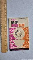 Front cover of vintage paperback book 'The Key to Tea-Cup Fortune Telling' showing colorful design with teacup illustration and ruler for size reference.