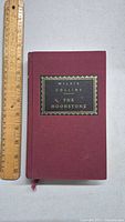 Front cover of the book showing maroon cloth cover with a decorative black and gold embossed label reading 'Willie Collins The Moonstone'. A wooden ruler next to the book measures about 7.5 inches height.