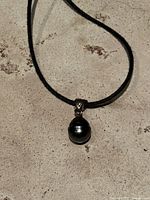 Full view of pendant necklace showing Tahitian black pearl on black cord against neutral background.