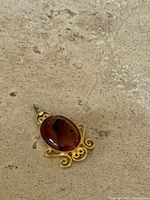 Front view of red-brown oval glass cabochon in gold tone Victorian style mount with scroll filigree work.