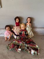 Four antique dolls displayed sitting upright against a wall on a stone surface; includes a small baby doll with curly hair in a pink crochet outfit, and three larger dolls in vintage-style dresses of red velvet and patterned fabric, silver and gold strapless fabric, and red and black plaid with lace trim.