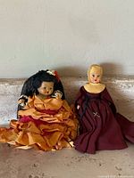 Photo showing two antique dolls, one dressed as a nun in maroon habit, and another in orange layered dress with black lace veil and floral headpiece.
