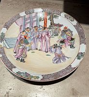 Full view of the round ceramic plate showing a painted scene of nine men in Qing Dynasty clothing around an architectural and garden setting, bordered with geometric and floral motifs.