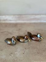 Three small metal duck sculptures with mixed metal finishes on a stone surface, shown from the front angle.
