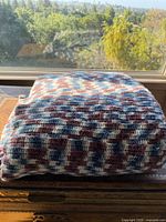 Folded large crochet blanket on a surface in front of a window, showing the colors and stitch pattern clearly under natural light.