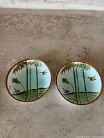 Top-down view of two round Kutani porcelain plates showing bamboo and bird garden scenes with red and gold rims on a textured surface.