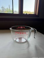 Clear vintage Pyrex glass measuring cup with red measurement markings visible on the side, including cups, ounces, and quarts, photographed near window light.