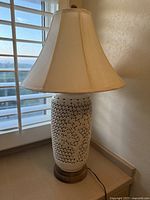 Full view of entire lamp showing reticulated white porcelain body, wooden base, cream fabric lampshade, and cord.