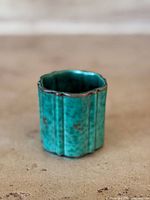 Front angle view of the small green turquoise glazed stoneware vase with silver inlay and scalloped shape on beige surface.
