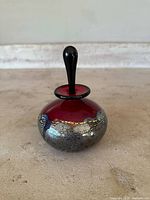 Multiple angled photos of a round red studio glass perfume bottle with black stopper and silver crackle design, signed and numbered by artist Michael Nourot.