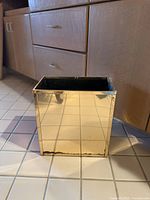 Rectangular gold-toned reflective waste bin with black interior lining, placed on tiled floor in front of wood cabinets. Shows wear along bottom edge.