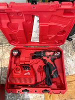 Open red hard plastic case containing Milwaukee cordless drill, battery charger, battery pack, and instruction manual.