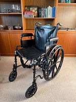 Front angle view of black Pride Mobility manual wheelchair positioned on a carpeted floor, showing padded seat, backrest, armrests, small front wheels and large rear wheels with push rims.
