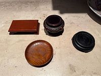 Four vintage Asian style wooden display stands on a light surface including a rectangular reddish-brown stand, a small round dark brown carved stand, a round light brown stand, and a black circular stand.