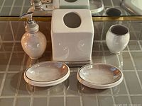 Photo showing five vintage bathroom accessory items arranged on a tiled surface in front of a mirror: a soap dispenser, square tissue box cover, toothbrush holder, and two soap dishes.