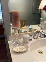 Photo of a bathroom sink area showing a clear glass candle holder with a pink candle, oval clear glass dish with gold geometric pattern, white ceramic container with a floral embossed lid, and a small metal cup, all arranged on the sink counter.