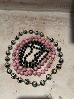 Overall view of the three coiled bead necklaces showing the black cloisonné beads with floral design, pink marbled beads, and small black beads on a concrete surface.