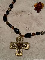Close-up photo of the gold-toned cross pendant on a mixed bead necklace showing detailed embossed texture and bead accents.
