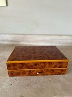 Closed burl wood rectangular jewelry box with visible wood grain patterns and lighter wood trim along edges and front drawer.
