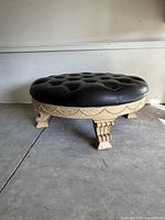 Side angle view of vintage black leather tufted ottoman with carved cream wooden base and legs, showing detail of tufting and leg carvings.