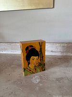 Side view of the rectangular wooden box showing the hand-painted face of an Asian woman with traditional hairstyle and clothing, orange background.