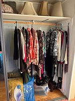 Closet rod with assorted women's garments hanging; shelf above holding six lamp shades.