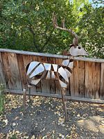 Front angle view of full life-size metal deer sculpture on ground near wooden fence