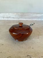 Single small vintage mahogany wood sugar bowl with turned lid and spoon resting beside or inside, placed on concrete surface.