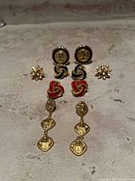 Photo showing seven pairs of vintage gold tone earrings on a textured surface including coin motif clip-ons, black and red enamel knots, floral studs, and textured dangles.