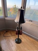 Full image of the table lamp showing leather shade and tassels with metal base on wooden floor near window.