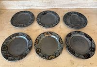 Six gunmetal grey ceramic plates arranged flat to show front surface and decorative rims with black, brown and green accents.