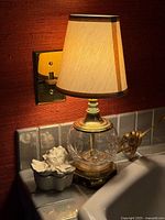 Photo of the vintage mini table lamp lit, showing cream fabric lampshade with gold trim, brass base and crystal ball body, alongside ceramic cherub jewelry box on a tiled surface.
