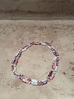 Image showing a vintage necklace made of polished stones in pink, lavender, and white colors laid out in a circular shape on a stone surface.